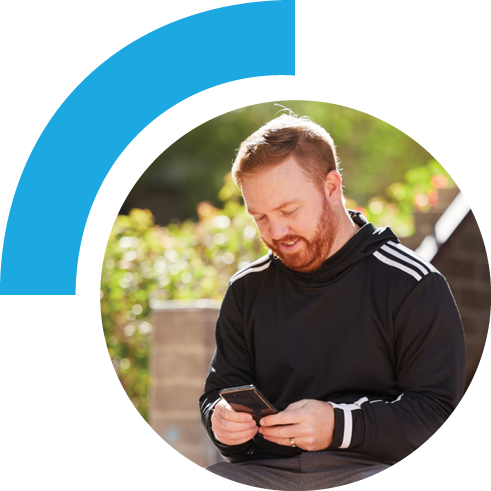Man sitting outside while on his phone. The man is pictured sitting front front of stairs with lush green plants in the background.