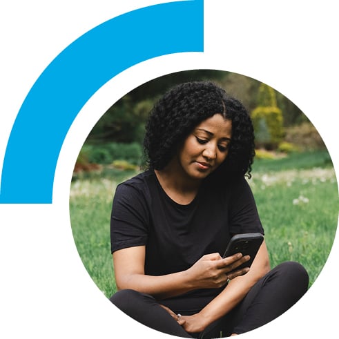 Woman on her phone while sitting in an open field of grass with a soccer ball on the side.
