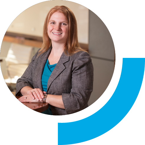 Woman in gray blazer smiling while at a desk with one hand over the other.