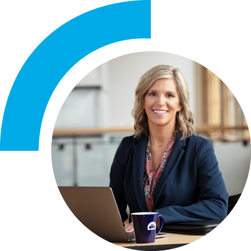 Woman in business attire sitting and smiling at a desk with laptop opened, cup of coffee, and notepad. The background setting is bright windows and brick walls emulating a professional feeling.