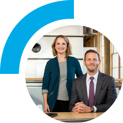 A woman in a teal cardigan stands smiling next to a seated man in a suit and tie at a desk with a laptop. Modern office with large windows in the background.