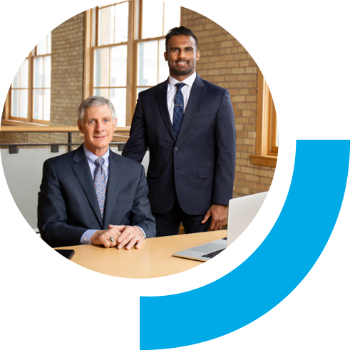 Two men in suits side by side with one sitting and the other standing. The background setting is bright windows and brick walls emulating a professional feeling.