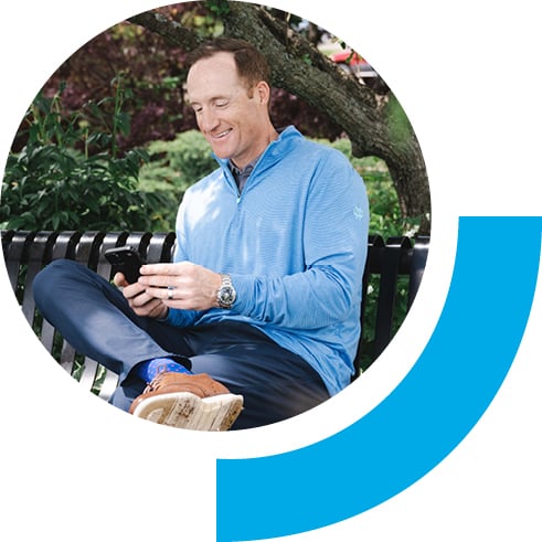 Middle-aged man sitting on a bench outside while on his phone. The background setting is green plants and small trees.