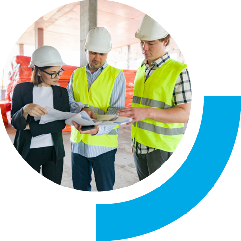 Two male workers in green vest with a woman in business casual clothing inside a building under construction. All three are wearing hard hats and appear to be chatting while holding papers.