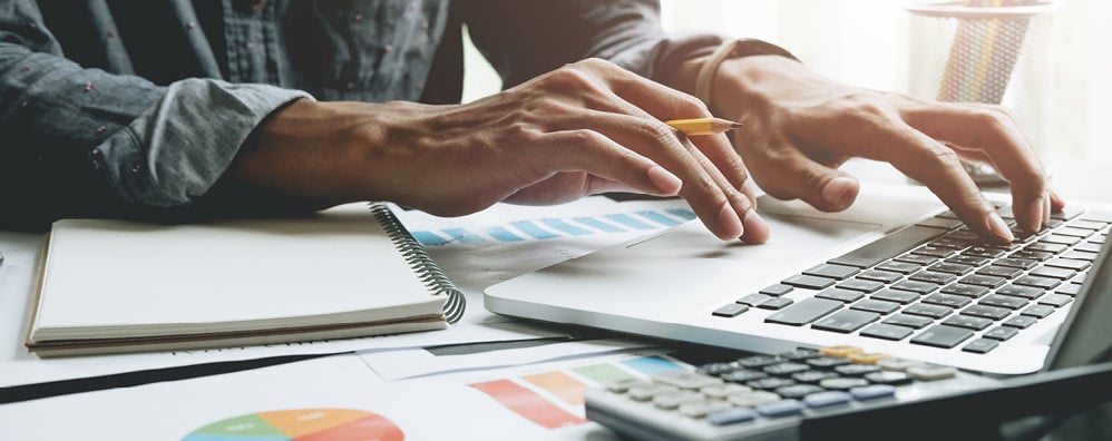 Person typing on laptop with pencil in hand and a calculator and notebook next to laptop