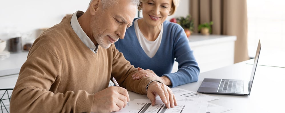 Elderly couple filling paperwork with laptop opened in their home