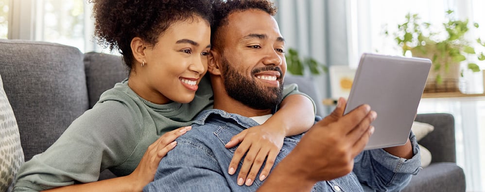 Young couple smiling and sitting on their couch staring at tablet