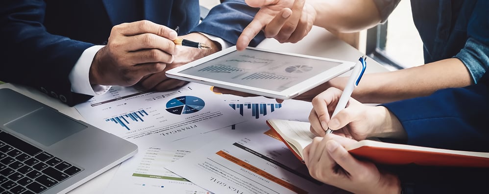 Three people review charts and graphs on documents and a tablet while one person takes notes. A laptop rests on the table.
