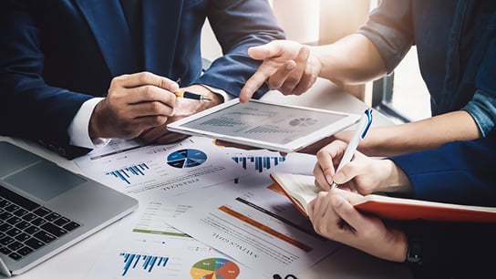 Three people review charts and graphs on documents and a tablet while one person takes notes. A laptop rests on the table.