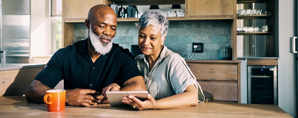 An elderly couple staring at a tablet while seated at a table in their home.