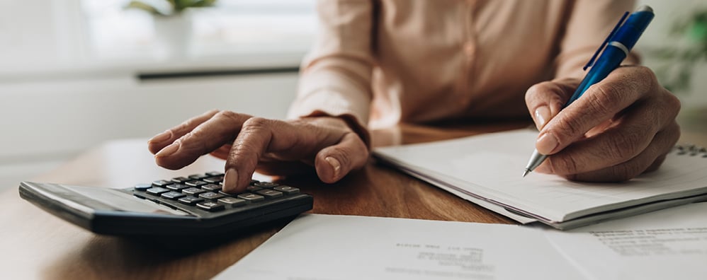 Person sitting at desk with pen in one hand on top of a notebook and papers, while touching a calculator in the other hand.