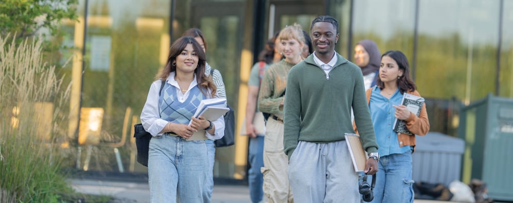 Multiple students walking out of a building carrying notebooks, books, and bags. The scenery is the exterior of an academic campus.