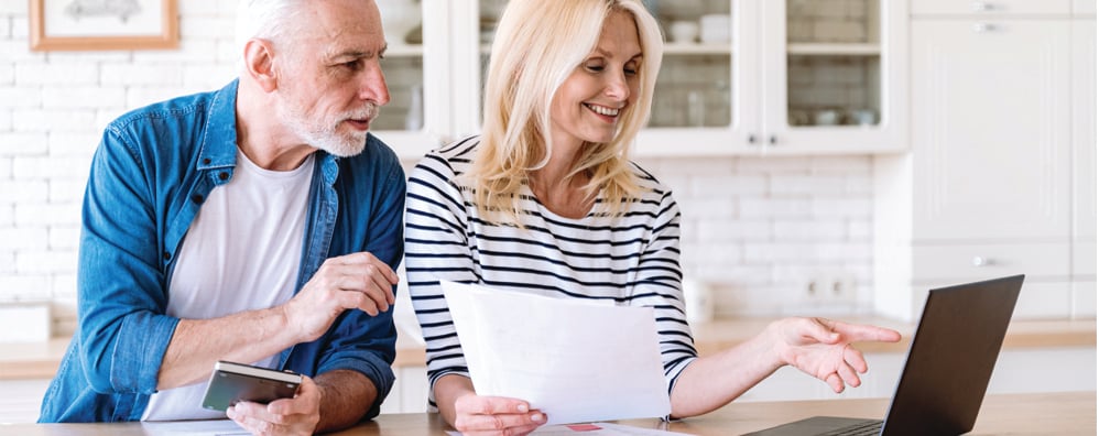 Elder couple looking at a laptop screen together. The couple can be seen standing over an island counter in a home environment.