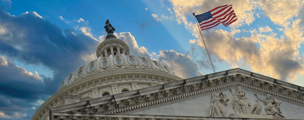 Government dome building under a sky with clouds and sunlight. An American flag waves atop the building.