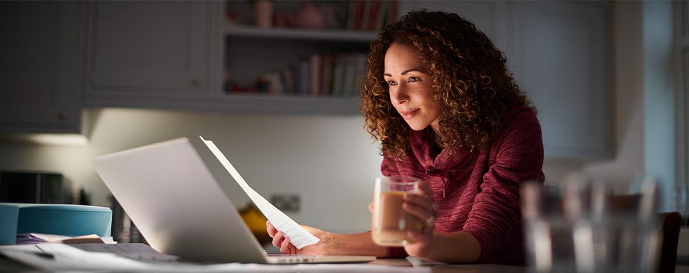 Woman with curly hair working on a laptop in a dimly lit kitchen, holding a mug. She appears focused and relaxed, creating a cozy atmosphere.