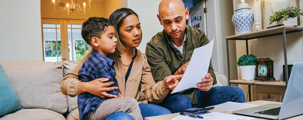 Adult couple sitting down in living room environment with son in mom's arms. The father is holding the paper as the mother points to it and the family appears to be discussing an important topic.