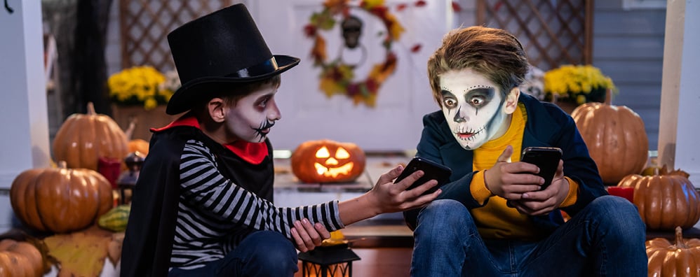 Two children in skeleton face paint sit on a porch surrounded by pumpkins. One wears a top hat and cape; both are holding phones, creating a festive Halloween atmosphere.