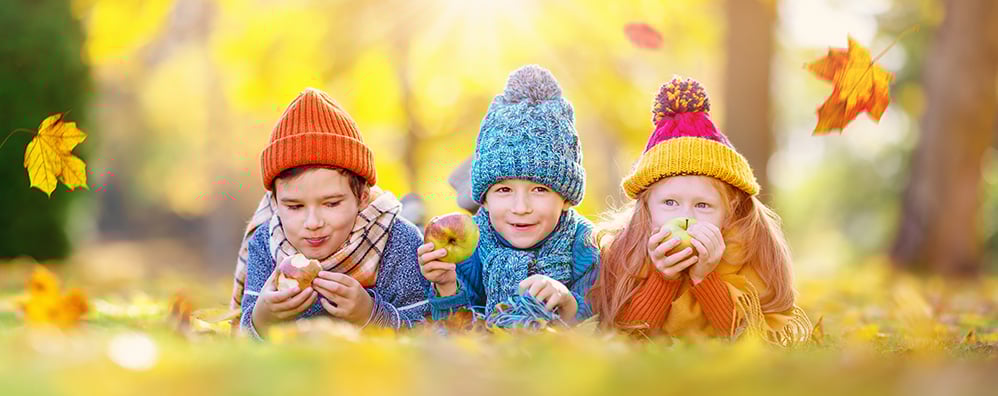 Three children in colorful knit hats and scarves lie on autumn leaves, joyfully eating apples. Golden leaves float around them under a sunny sky.