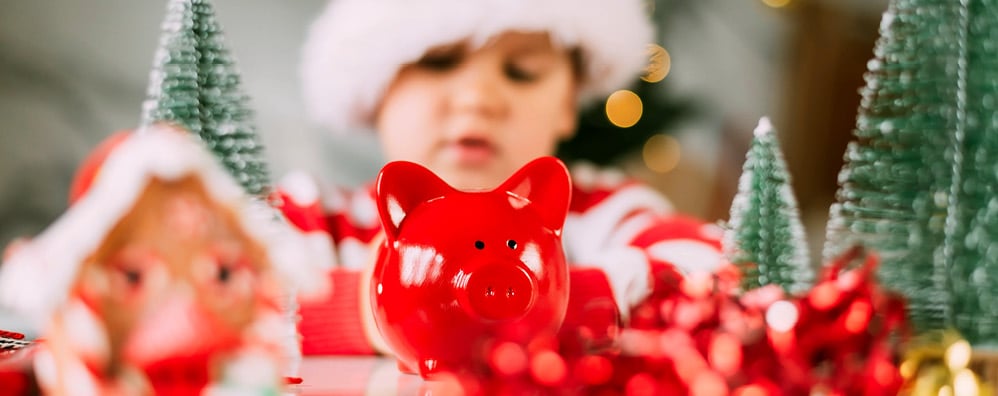 A small, red piggy bank sits in front of a child with a Santa hat and striped, red shirt on a table surrounded by Christmas decorations.