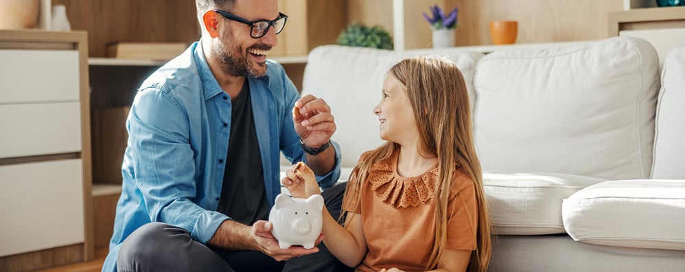A man and girl sit smiling on a living room floor with a piggy bank. The man holds the piggy bank while the girl prepares to deposit a coin, highlighting saving money.