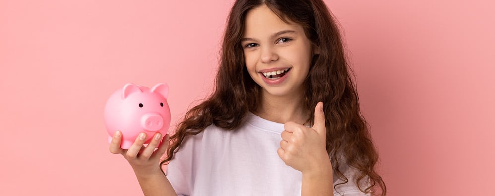 A smiling girl holds a pink piggy bank in one hand, giving a thumbs-up with the other. She stands against a matching pink background, wearing a white shirt.