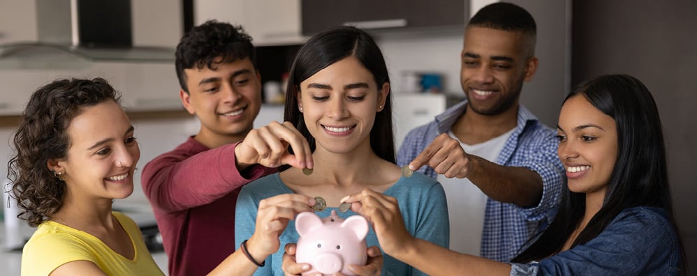 A group of five, smiling friends - one person is holding a pink piggy bank while the other 4  place coins into the piggy bank.