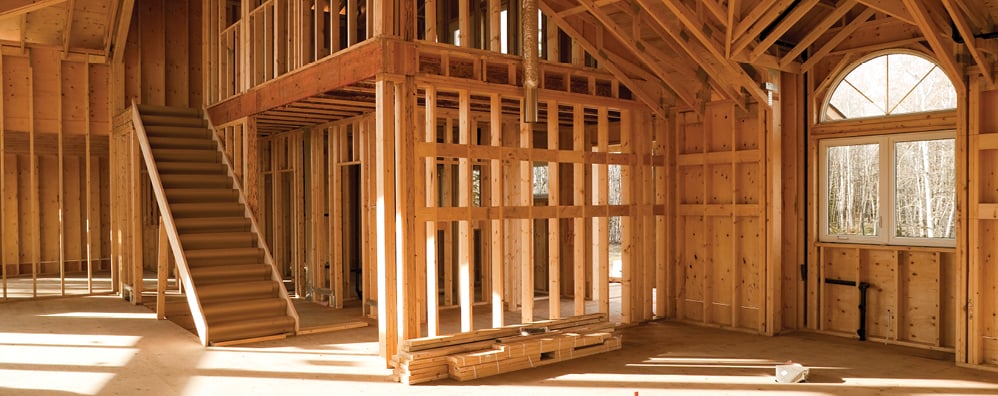 Interior of a house under construction with exposed wooden beams, partial framing, and a staircase in a sunny environment.