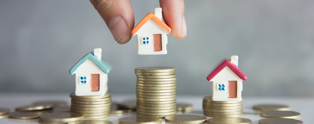 A hand carefully positions a tiny house atop stacks of coins