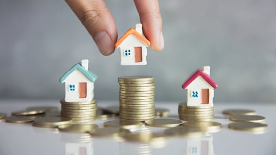 A hand carefully positions a tiny house atop stacks of coins