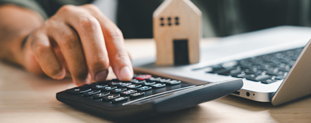 Person using calculator next to a laptop that has a miniature house figure on top.