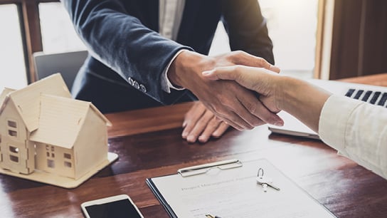 Handshake over a desk with a wooden house sculpture on the side.