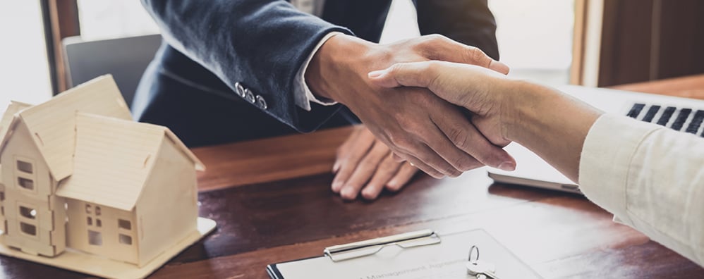Handshake over a desk with a wooden house sculpture on the side.