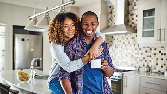A joyful couple in a modern kitchen, with one hugging the other from behind. They smile warmly, holding a coffee mug, conveying happiness and togetherness.