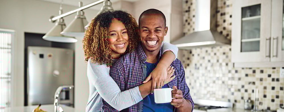 A joyful couple in a modern kitchen, with one hugging the other from behind. They smile warmly, holding a coffee mug, conveying happiness and togetherness.