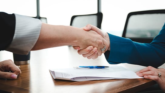 Two business people shaking hands at a desk on top of papers and pen.