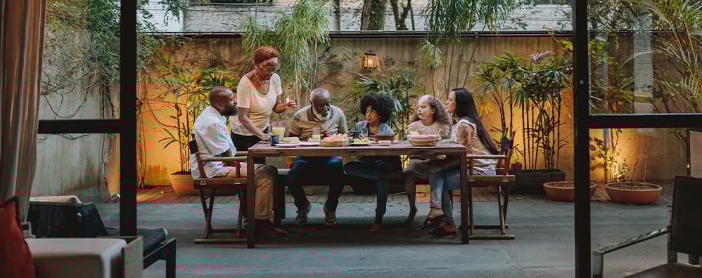 Multi-generational family having dinner outside in a cozy garden patio with warm lighting and lush plants creating a relaxed mood.