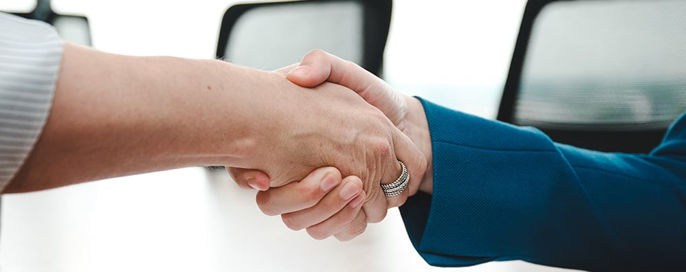 Two business people shaking hands at a desk on top of papers and pen.