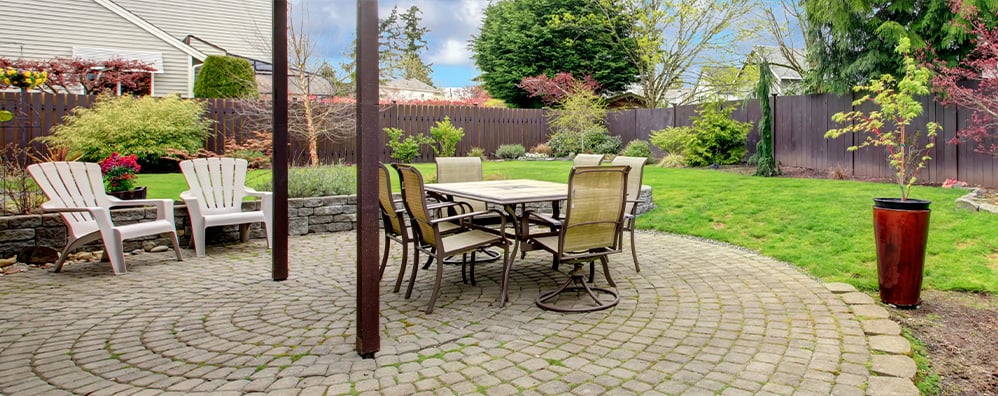 A tranquil backyard patio with a round stone pavement, featuring a dining table and six chairs. Two white chairs sit to the side. The garden is lush with green grass, various plants, and a large planter, enclosed by a brown wooden fence under a clear blue sky.