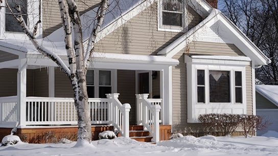 A cozy house in winter with snow-covered ground and icicles hanging from the porch. A bare tree stands beside the house under a clear blue sky.