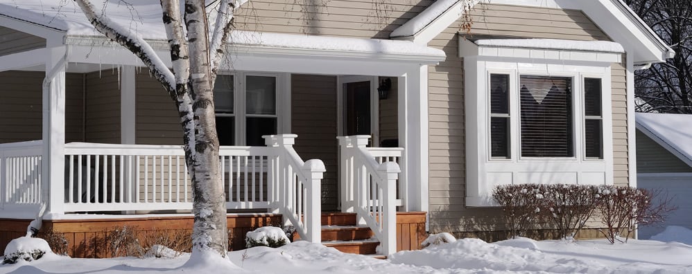 A cozy house in winter with snow-covered ground and icicles hanging from the porch. A bare tree stands beside the house under a clear blue sky.