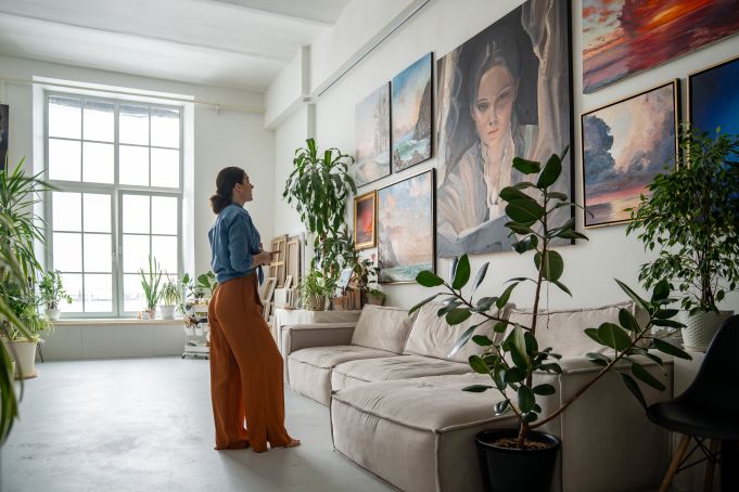 A woman in a bright, plant-filled art studio gazes at paintings on a wall. The room feels serene and creative, with natural light from a large window.