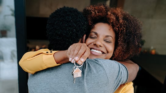 A woman with curly hair happily hugs another person, holding a key with a house-shaped keychain. 