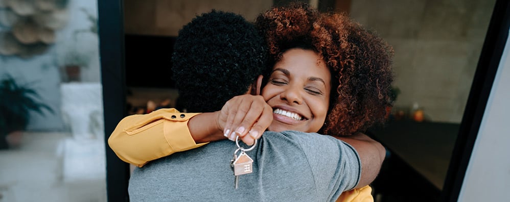 A woman with curly hair happily hugs another person, holding a key with a house-shaped keychain. 