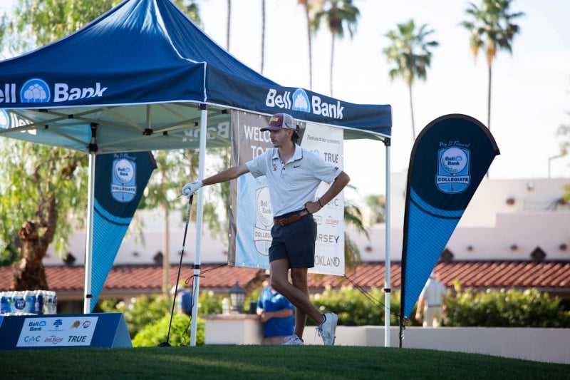 Man standing with their golf club with the Bell Bank tent and sponsor signs in the background