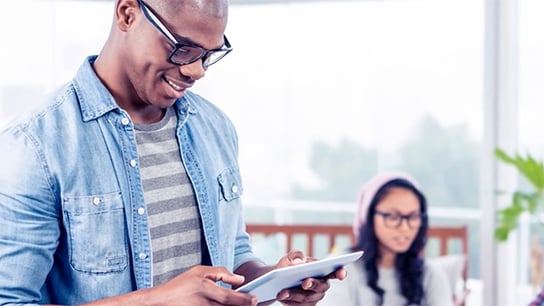 A man smiling and looking at a tablet with a women in the background smiling