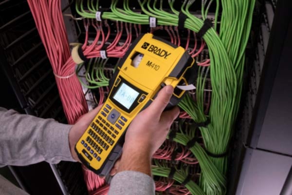 A person&#x27;s hand holds a black and yellow portable Brady M410 label printer in a data center environment. The printer has a label partially dispensed, showing printed text intended for cable identification.