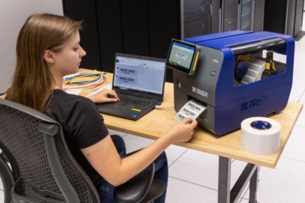 A woman using a blue Brady i7500 cable printer/applicator to automatically apply a clear, self-laminating label to a label server racks.