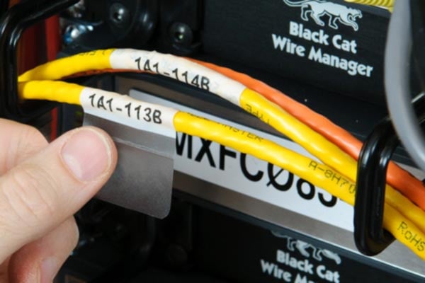 A person's hands are shown inspecting a clear, self-laminating label to a single network cable in a data center. The label has printed text on it and is being wrapped around the cable, with the clear tail overlapping the printed section for protection.