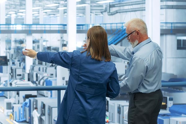 Two figures on a balcony survey a manufacturing floor. 