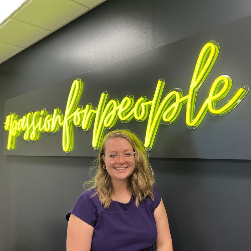 Brunel Account Coordinator Abbi DeBeck smiles and poses in front of a neon yellow sign that reads #passionforpeople